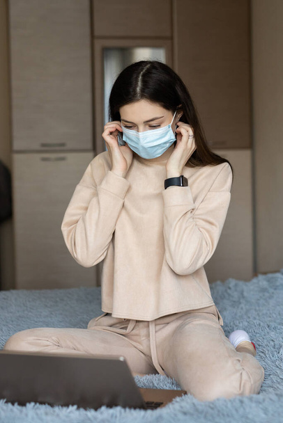 junge Studentin in schützender blauer medizinischer Maske auf dem Bett, Arbeit mit einem Laptop-Notebook, Online-Bildungseinkauf, Arbeit zu Hause während der Quarantäne covid-19 - Foto, Bild