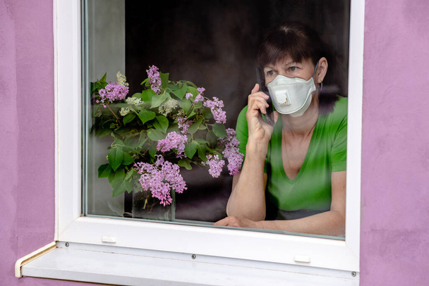 A sad older woman in a protective mask is bored, looks out the window and talks on the phone with the children. The concept of coronavirus quarantine stay at home and social distance. - Foto, imagen