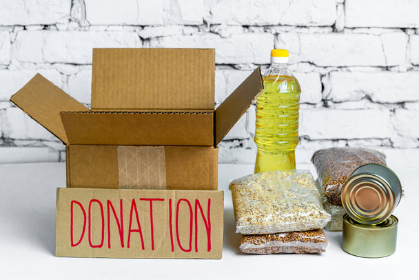 Food in cardboard donation box on a white background. Anti-crisis stock of essential goods for period of quarantine isolation. Food delivery, coronavirus. The shortage of food. Sign with text. - Foto, Imagen