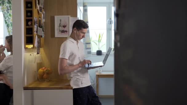 A young father works on a laptop in the kitchen while the mother and child play in the next room, a video conference from home - Záběry, video