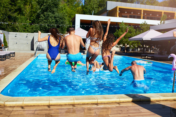 Happy friends jumping and splashing in swimming pool with inflatable floats in luxury resort. Young people in swimwear having party in private holiday villa on sunny day. Hot girls jump into water. - Photo, Image