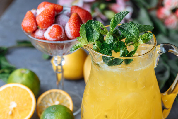 Summer fresh cold drink beverages. Ice Lemonade in the jug and lemons and orange with mint on the table outdoor. Orange lemonade in a jug. - Foto, Bild