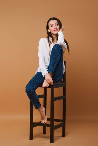 Lovely girl in white shirt and jeans sitting on tall wooden chair in studio - Foto, Bild