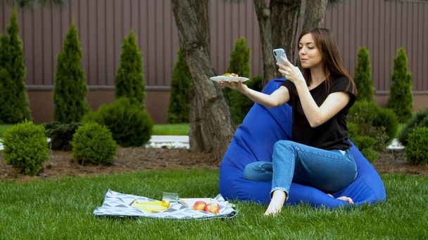 Νεαρή γυναίκα φωτογραφίζει φρούτα και σάντουιτς στον κήπο - Φωτογραφία, εικόνα
