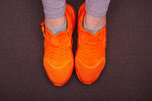 Gambe femminili in pantaloni della tuta grigi, leggings in scarpe da ginnastica arancione brillante per lo sport, jogging, fitness con suole alte su Karimat grigio. Gambe femminili in scarpe da ginnastica arancioni su un tappeto grigio per yoga, sport, turismo
 - Foto, immagini