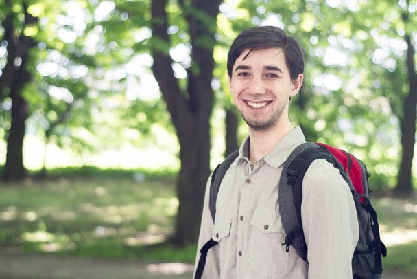 Красива молода брюнетка в сірій сорочці з рюкзаком стоїть в парку і посміхається. Концепція подорожі з простором копіювання
. - Фото, зображення