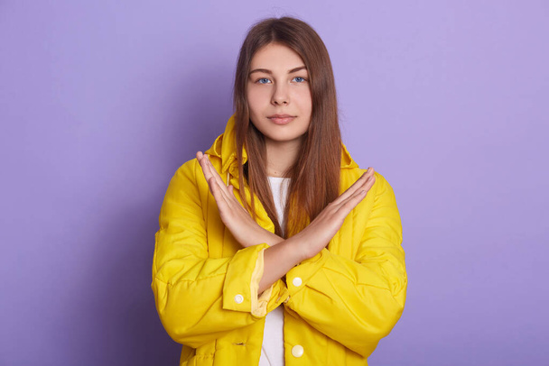 Indoor shot of serious or sad woman posing isolated over lilac studio background, lady wearing yellow jacket, crossing hands, female showing stop gesture. Body language, people, lifestyle concept. - Φωτογραφία, εικόνα