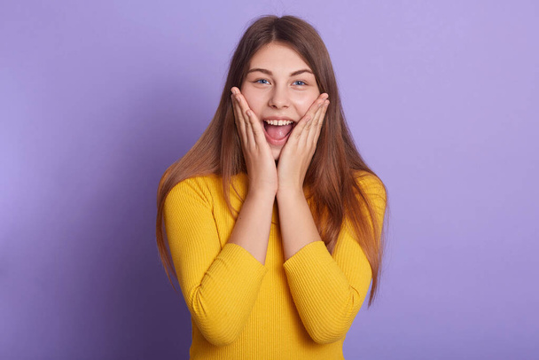 Studio shot of attractive female yelling happily, keeping hands on her cheeks, lady with long hair dresses yellow shirt, standing against lilac background, has positive emotions and happy expressions. - Valokuva, kuva