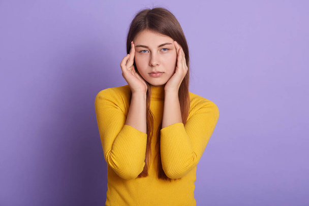Closeup portrait of unhappy woman with serious facial expression, looking directly at camera, keeping fingers on her temples, dresses casually, posing isolated over lilac studio background. - Photo, image