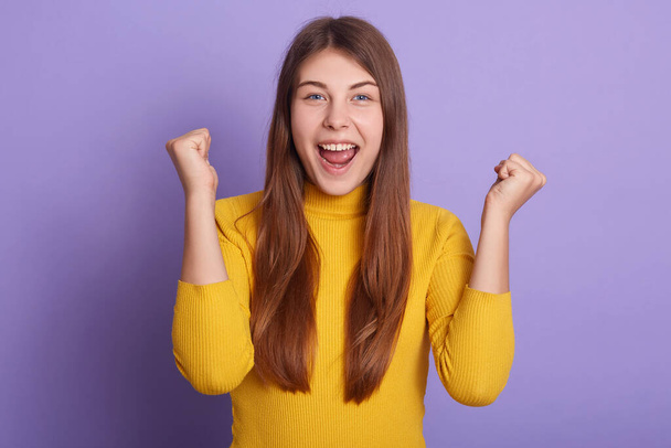 Closeup portrait of happy woman clenching fists and celebrating something happily, wearing casual attire, keeping mouth opened, yelling something, posing isolated over lilac studio background. - Photo, Image