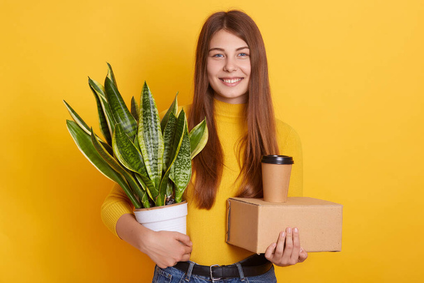 Indoor shot of happy adorable Caucasian woman wearing yellow casual shirt, lady holding carton box, take away coffee and flowerpot with Sansevieria, looking directly at camera, being in good mood. - Φωτογραφία, εικόνα