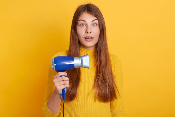 Closeup portrait of shocked female with long beautiful hair, holding hair dryer in hands, looking at camera with big eyes, being surprised, wearing casual closing, posing against studio wall. - Photo, Image