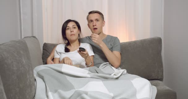 Young couple wrapped in a plaid eating popcorn and watching tv while sitting on the sofa at home. Leisure and recreation concept. - Felvétel, videó