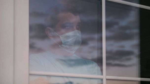 家庭用隔離,コロナウイルス病のための自己分離, COVID-19.窓の近くの医療マスクの男。検疫中の退屈、悲しい、うつ病や自殺気分。ガラス越しの眺め. - 写真・画像