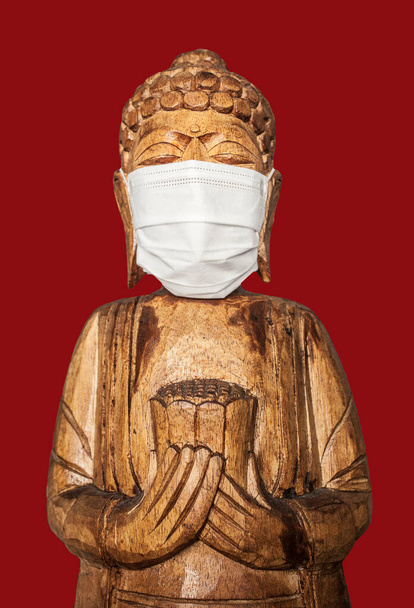 Boeddha houten standbeeld met gezichtsmasker. Geïsoleerd boven rood. Covid-19-concept - Foto, afbeelding