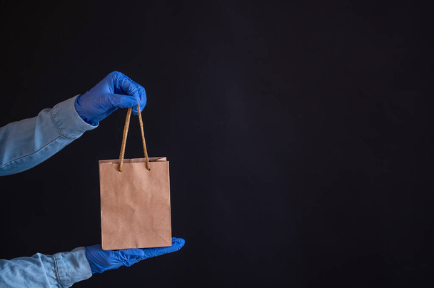 Female hands in blue gloves holds a brown paper bag with handles on a black background. Safe food delivery to your home. A courier in a denim shirt holds out a craft cardboard bag to a customer. - Foto, afbeelding