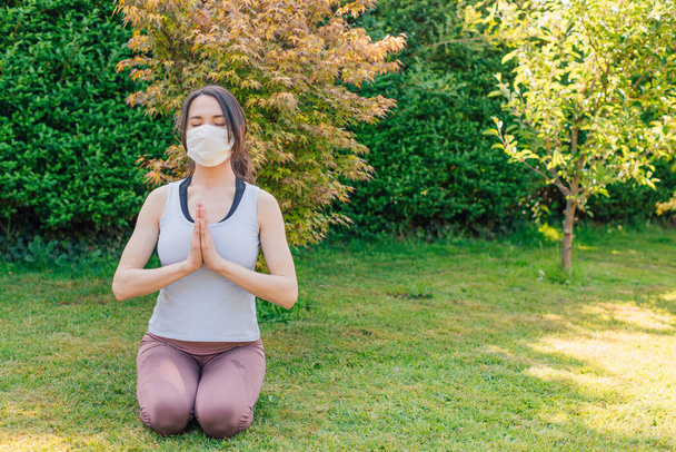 Young woman meditating outdoors in a park or garden with surgical facemask - concept of making yoga and fitness exercises during or after coronavirus outbreak - Foto, Bild