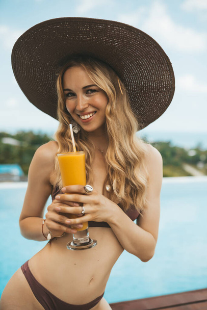 Smiling blonde woman in summer hat sitting at swimming pool and drinking mango shake - Foto, imagen