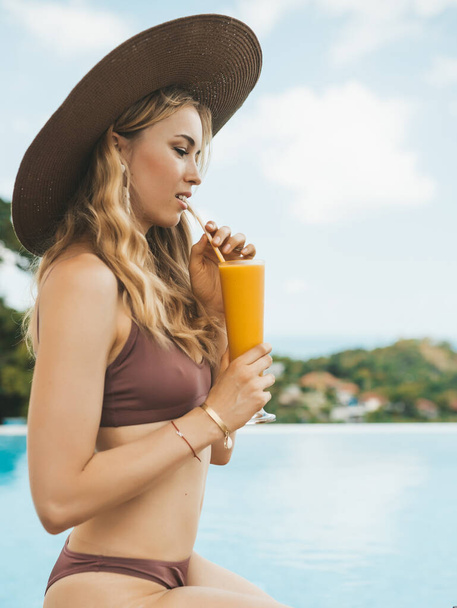 Smiling blonde woman in summer hat sitting at swimming pool and drinking mango shake - 写真・画像