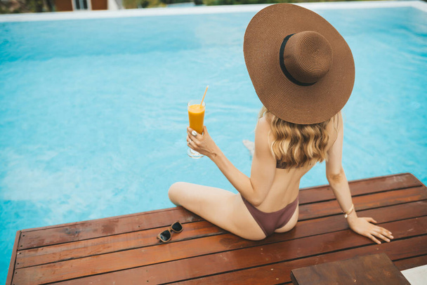 Smiling blonde woman in summer hat sitting at swimming pool and drinking mango shake - Foto, immagini