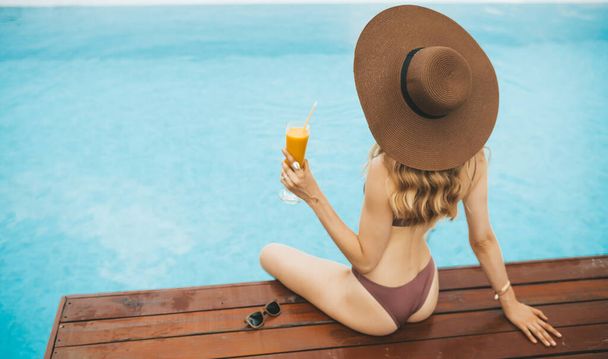 Lächelnde blonde Frau mit Sommermütze sitzt am Swimmingpool und trinkt Mango Shake - Foto, Bild