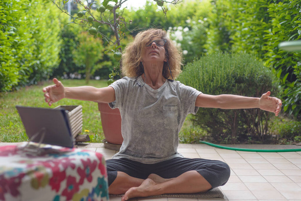 Gözlüklü olgun bir kadın tabletle yoga pozisyonunda, dışarıda yeşil bahçede meditasyon yapıyor. Covid-19 karantinası evde kalacak. Kilitleme çözümü. - Fotoğraf, Görsel