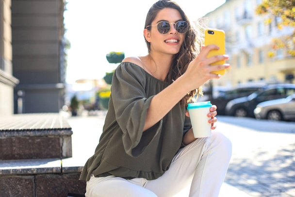 Χαμογελώντας όμορφη μελαχρινή γυναίκα με τα πόδια σε εξωτερικούς χώρους από το δρόμο, να πάρει selfie από το κινητό τηλέφωνο, πίνοντας καφέ - Φωτογραφία, εικόνα