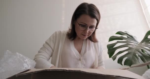 Mature woman unpacks cardboard box on table at home, in office - Video, Çekim