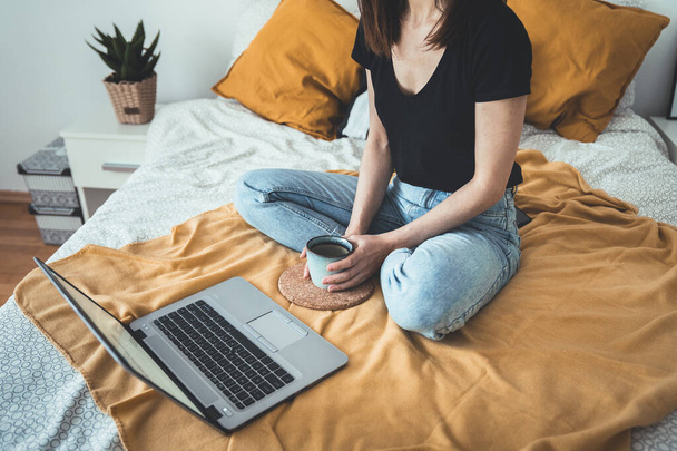 若い女性は、寝室でラップトップコンピュータを使用してホットコーヒーや紅茶のカップをリラックスして飲みます.コミュニケーションとテクノロジーの概念 - 写真・画像