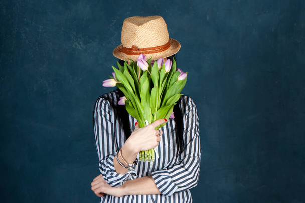 Trendy beautiful girl, brunette in summer straw hat and striped shirt, with a bouquet of tulips flowers, covered her face with a bouquet, the theme of summer and spring.  - Fotó, kép