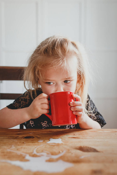 cute little girl drinks milk from a red mug at home - Fotoğraf, Görsel