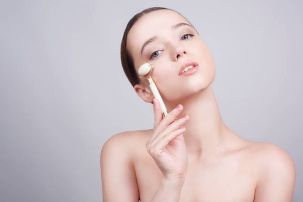 Beauty face care. Woman doing face massage with white jade facial rollers for spa skin care treatment at home. Girl model using natural massager tool portrait on white background - Photo, Image
