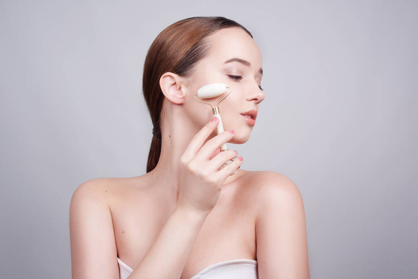 Portrait photo of a young woman looking relaxed use while using a natural white quartz face roller. Beauty face care. Woman doing face massage with jade facial rollers for spa skin care treatment at home.  - Zdjęcie, obraz