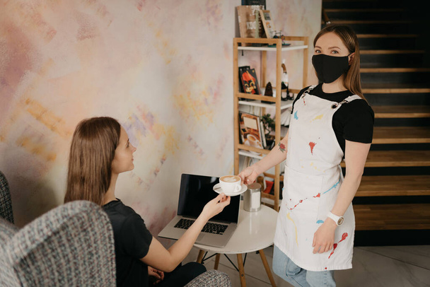 A female barista in a black face mask serving a girl a cappuccino in a coffee shop. A girl with long hair working remotely on a laptop keeps social distance ordered a coffee in a cafe.    - Photo, Image