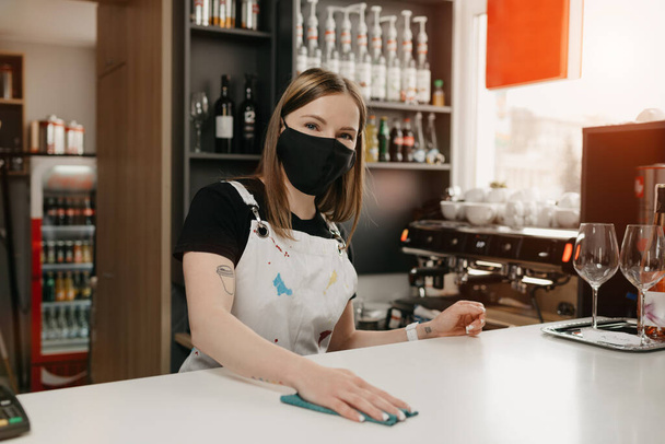 Una barista con tatuajes en una máscara médica negra sonríe y limpia el mostrador de la barra en una cafetería. Una hermosa camarera desinfectando un café
. - Foto, imagen