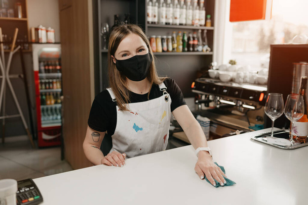 A happy female barista with tattoos in a medical black face mask smiles and cleaning the bar counter in a coffee shop. A beautiful waitress disinfecting a cafe while waiting for clients. - Photo, Image