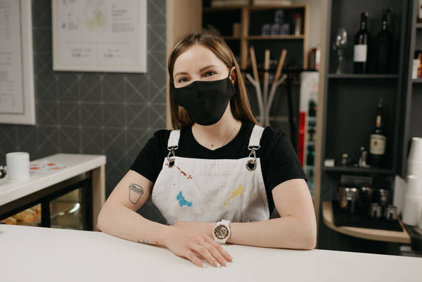 A happy female barista with tattoos in a medical black face mask smiles and waiting for clients in the coffee shop. A beautiful female owner of the cafe poses with arms crossed behind the bar.  - Fotografie, Obrázek