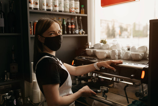 医療用の黒いフェイスマスクをしたタトゥーのある幸せな女性バリスタが、カフェでコーヒーを淹れます。コーヒーマシンの近くにポーズをとるウェイトレス.  - 写真・画像