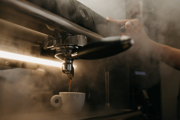 Μια κοντινή φωτογραφία μιας επαγγελματικής καφετιέρας. Ένας μπάρμαν χύνει έναν εσπρέσο από μια καφετιέρα σε ένα λευκό φλιτζάνι. Ατμός γύρω. - Φωτογραφία, εικόνα