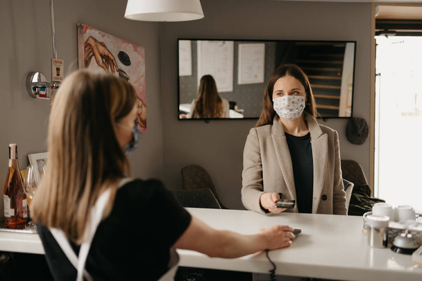 A girl who wears a medical face mask uses a cell phone to pay by contactless NFC technology. A female barista in a face mask holds out to a client a terminal for paying. - Zdjęcie, obraz