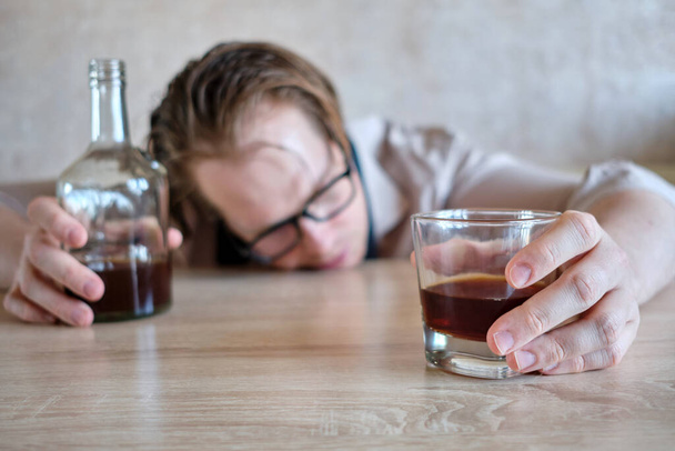 Ein Mann liegt mit dem Gesicht auf einem Tisch mit einem Glas und einer Flasche Alkohol. Nahaufnahme. - Foto, Bild