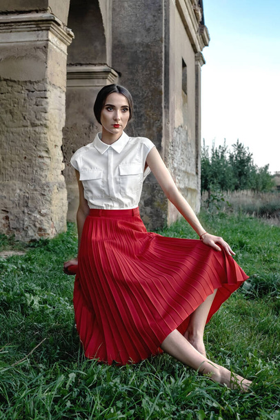 Beauty Fashion Model Girl with brown hair against the background of an old, abandoned building, in a stylish red skirt and shirt. Sexy woman portrait with perfect make up and fashion clothes. - Photo, image