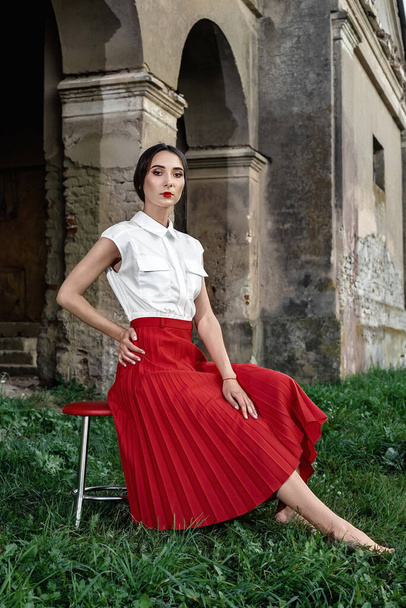 Beauty Fashion Model Girl with brown hair against the background of an old, abandoned building, in a stylish red skirt and shirt. Sexy woman portrait with perfect make up and fashion clothes. - Photo, image