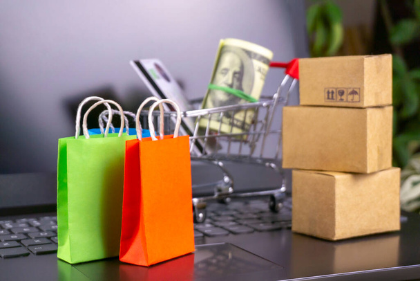 online winkelen concept. kleine winkelwagen, dozen, kleurrijke tassen, bankkaart, dollars staan op laptop - Foto, afbeelding