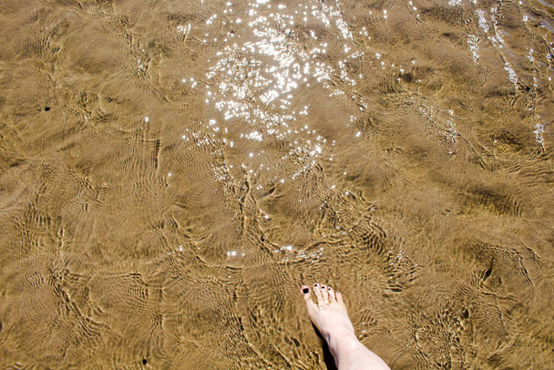 O pé da menina com esmalte preto vem na areia coberta com uma camada transparente de água que brilha ao sol
 - Foto, Imagem
