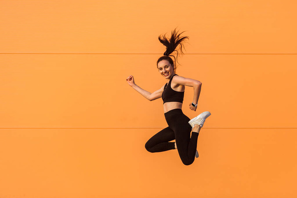 Happy lively vivid girl with fit body in tight sportswear jumping in air, flying and smiling full of energy, healthy sport lifestyle. studio shot, isolated on orange background, advertising area - Foto, afbeelding