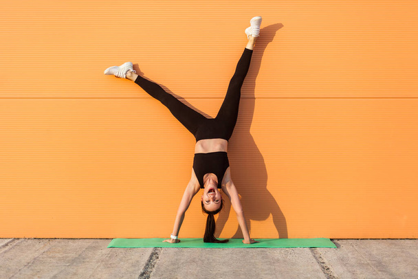 Overjoyed excited girl with perfect athletic body in tight sportswear doing yoga handstand pose against wall and laughing, shouting from happiness. Gymnastics for body balance outdoor workouts - 写真・画像