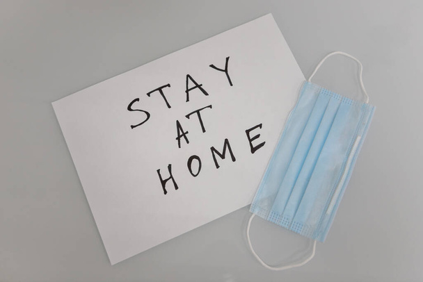 Lékařská ochranná maska na stole u bílého papírového listu s nápisem "Zůstaňte doma". Výzva k sebeizolaci během karantény během pandemie koronaviru. - Fotografie, Obrázek