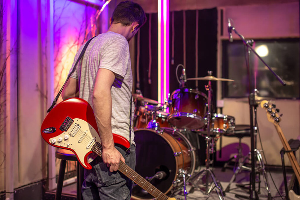 Um homem toca uma guitarra elétrica em um estúdio de gravação. Uma sala de ensaios para músicos com um kit de bateria em segundo plano. O conceito de criatividade musical
. - Foto, Imagem
