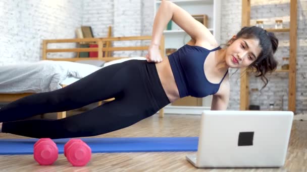 20s young Asian woman in sportswear doing stretching exercise while watching fitness training class on computer laptop online. Healthy girl exercising in living room with sofa couch in the background - Filmati, video